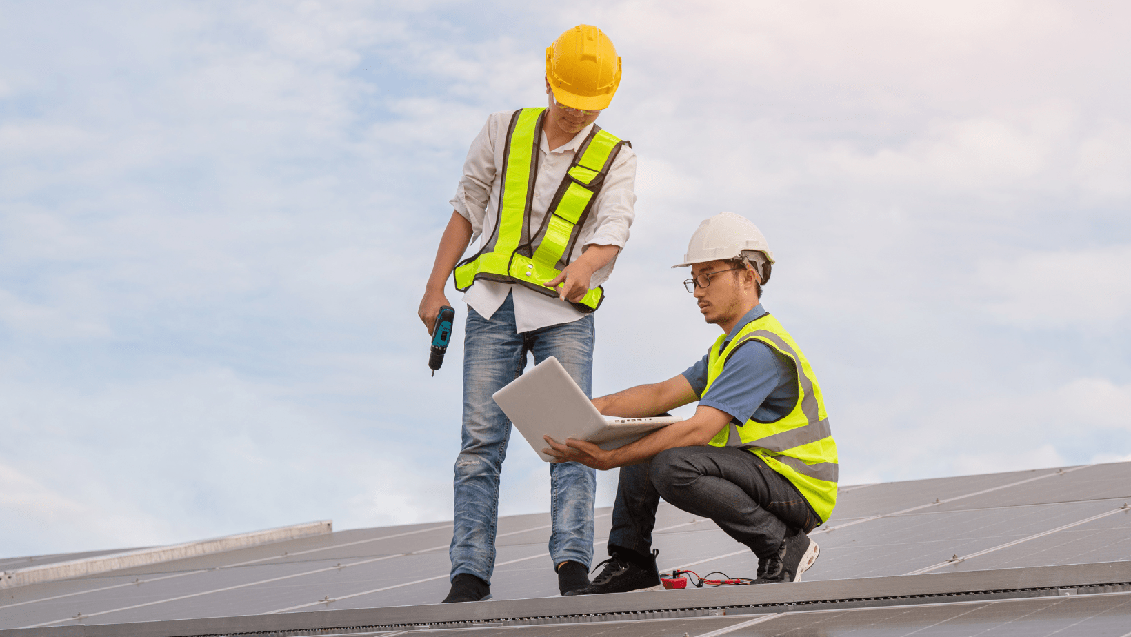 Two men in safety vests on a roof, collaborating while examining a laptop screen together.