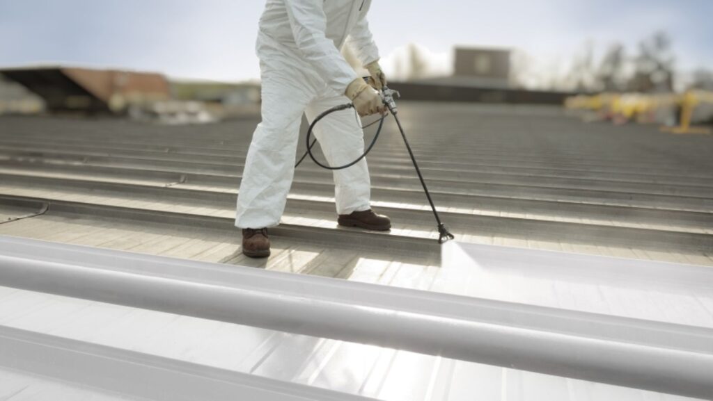 A man in white protective gear sprays a metal roof with a paint sprayer